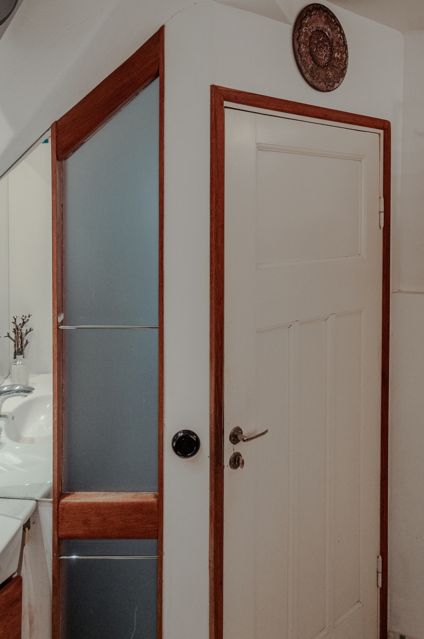 Badrumsinteriör visande vit dörr med eklister, ljusgenomsläpp i frostat glas med eklister, spegelvägg och vita väggar.