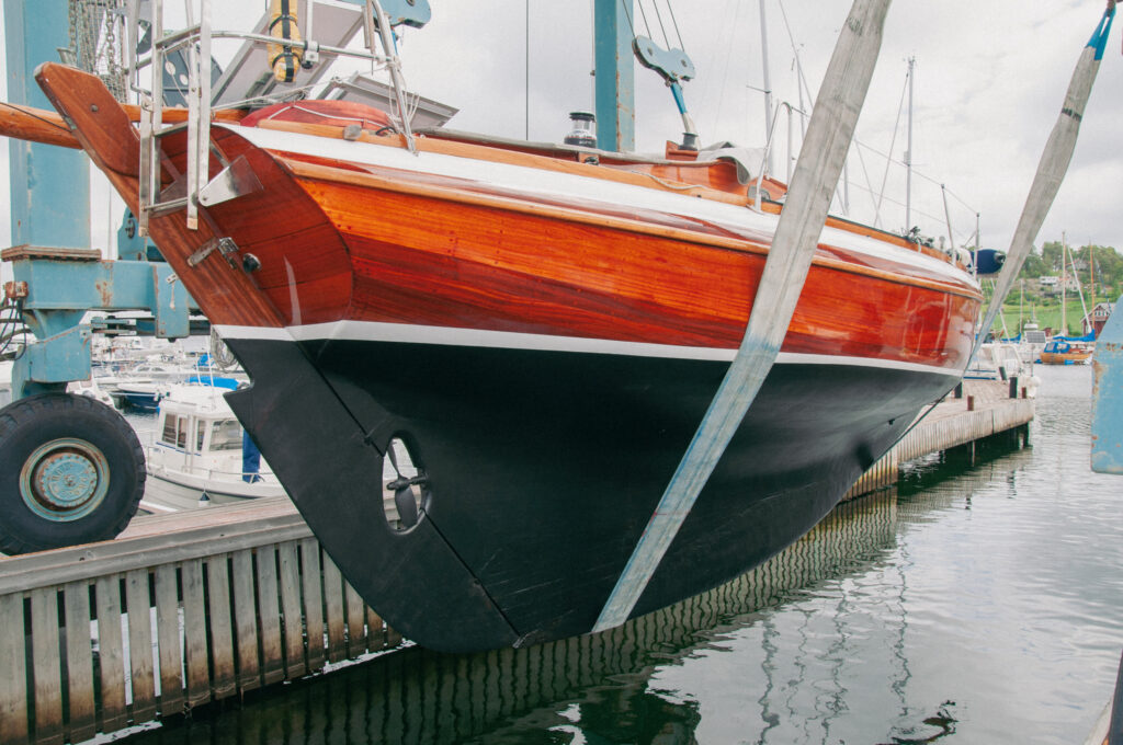 Blänkande mahognysegelbåt på väg att sjösättas - hänger strax ovanför vattenytan i sjösättningskranens lyftband